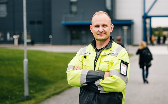 Mies keltaisessa työtakissa katsoo kameraan, taustalla teollisuusrakennus ja nurmikkoa.